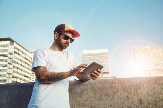 Low angle shot of a young man wearing a blank white t-shirt, baseball cap and sunglasses with a tablet in a city landscape with sun reflections