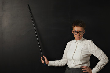Angry attractive woman teacher looks at camera and shows on black chalk board behind her with folding pointer