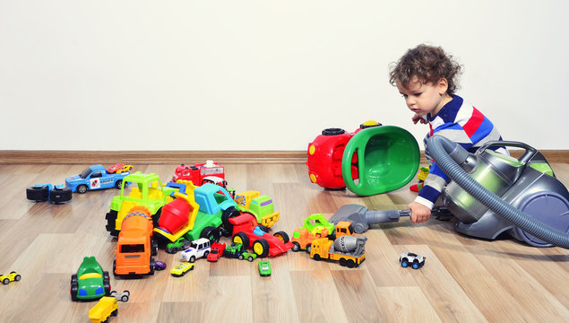 Mother Cleaning The Messy Room Full Of Toys. Close Up On Woman's Hands Cleaning With A Vacuum Cleaner. Upset Kid Playing With Many Toys On The Floor