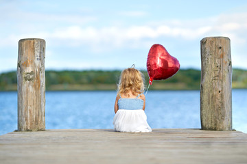 kleines Mädchen sitzt am See mit roten Herzluftballon