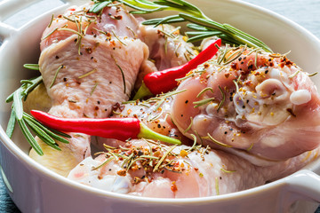 Raw chicken legs (drumsticks) in white baking dish, closeup. Black stone background, small chili pepper, rosemary and spices