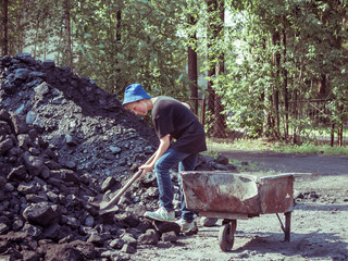 Toned image dirty little boy with a shovel in his hands which stands beside iron trolley and digging coal against the backdrop of a huge pile of coal with a fence and trees