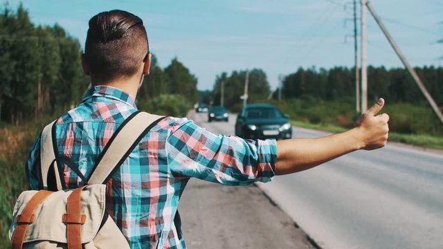 Back side of boy with backpack hitchhiking at road in summer sunny day. Cars. Countryside.
