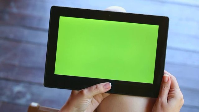 Young woman sitting over wooden background browsing internet and watching video on tablet PC with green screen. Point of view video footage of hands of anonymous woman and modern touchpad.