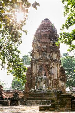 Buddha statue sitting position at front of pagoda under sun light surround by trees and ancient ruins of Wat Phra Mahathat temple in Phra Nakhon Si Ayutthaya Historical Park, Thailand