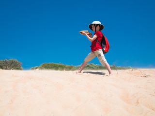 Little boy with a backpack and a crossbow in his hands stands on the golden sands and takes aim against the clear blue sky