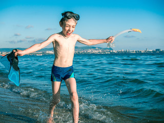 Toned image of a little boy in a mask for diving who holds in his hand flippers and snorkel and runs along the coast on the background of waves splashing and blue sky