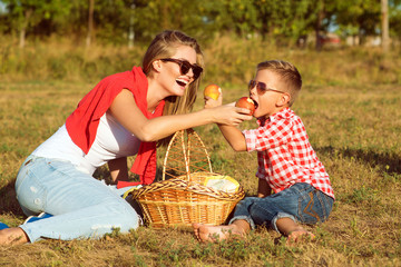 Young gorgeous mother and her little son in sunglasses and jeans having picnic in the countryside. They are sitting on the grass, laughing and feeding each other. Happy family concept. Outdoor shot