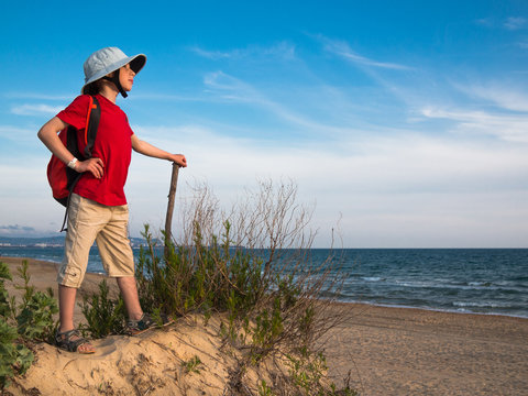 Toned image of a little boy with a backpack and a hat that holds the stick and standing on a sandy hill against the background of the calm sea and clear sky