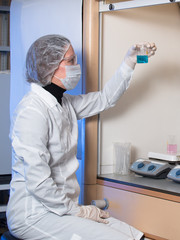Adult woman protective glasses and medical mask holding in his hand glass flask with blue liquid and in the other hand she is holding a syringe against the background of medical equipment for research