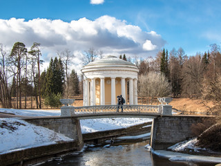 Toned image kissing couple standing on a bridge next to the house temple of Friendship in Pavlovsk Park (1780) in spring on the banks of the river Slavyanka against a blue sky with clouds
