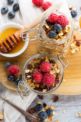 Fresh granola, muesli with berries, honey in a glass jars on a white wooden background, top view