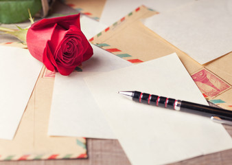 Vintage envelopes, red rose and sheets of paper scattered on the wooden table for writing romantic...