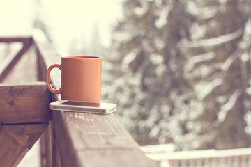 A cup with a hot drink and mobil phone on the background of the winter forest