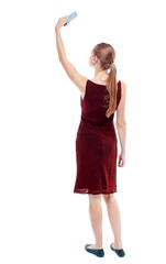 back view of standing young beautiful woman using a mobile phone. Isolated over white background. The girl in the maroon sleeveless dress makes selfie.