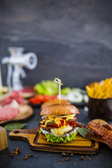 Ingredients for preparation burgers. Raw ground beef hamburgers on wooden board with meat grinder onion, greens, pickles, tomatoes, sauce, black stone background.

