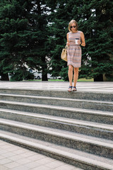 Portrait of young brunette woman in dress and sunglasses walking down the steps with coffee to go against the green trees