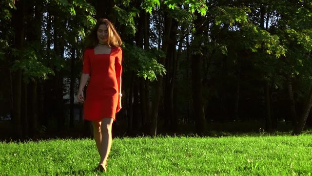 Slender brunette girl in red dress walking on the grass in the park. Slow motion video, 120 fps