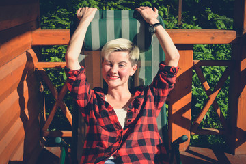 Obraz na płótnie Canvas Fashionable portrait of a young beautiful woman in red lumberjack shirt, jeans, white t-shirt and sneakers. Trendy blonde hipster girl having fun on a terrace of a rich wooden house on a sunny day.