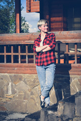 Fashionable portrait of a young beautiful woman in red lumberjack shirt, jeans, white t-shirt and sneakers. Trendy blonde hipster girl having fun on a terrace of a rich wooden house on a sunny day.