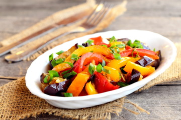 Vegetarian stew on a plate and on wooden table. Steamed aubergine, red and orange peppers and green onions. Diet dish. Vegetable stew dish. Closeup
