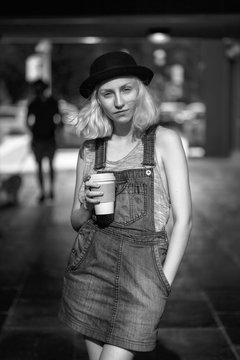 Black and white portrait of Caucasian teenage young blonde alternative model girl woman in tshirt, jeans romper looking in camera holding cup of coffee