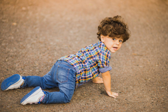 little cute toddler boy crawling outside. Caucasian child dressed in casual clothing: blue denim jeans, shirt and sneakers. portrait of kid with beautiful dark curly hair. Horizontal color image.