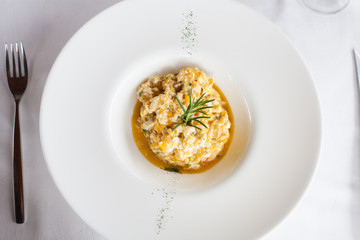 aerial view of elegant white circular bowl plate with scrambled egg with cod and fish, decorated with green rosemary leave, on white tablecloth in restaurant
