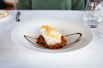elegant white circular bowl plate with hake fish cooked with typical vegetables named “pisto”, on white tablecloth in restaurant
