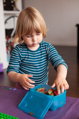 blonde two years old child with striped blue and white sweater inside home storing  plasticine in blue plastic box on purple table 

