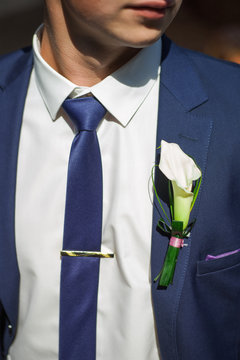 Details of male wedding clothes. Close up of white flower corsage. Beautiful boutonniere pinned on man in blue suit, white shirt and blue tie. Groom or graduate