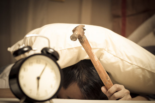 sleeping asian young male disturbed by alarm clock early morning on bed & holding hammer in hand