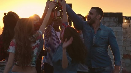 Group of multi-ethnic young people toasting with beer bottles and dancing to the music played by dj at urban rooftop party at sunset - Powered by Adobe
