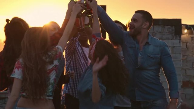 Group of multi-ethnic young people toasting with beer bottles and dancing to the music played by dj at urban rooftop party at sunset