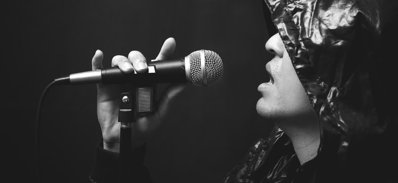 asian handsome male rocker artist in black leather hood jacket singing with dynamic microphone on dark background, bw filter