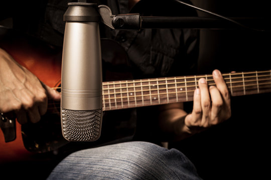 male musician playing acoustic guitar behind condenser microphone in recording studio