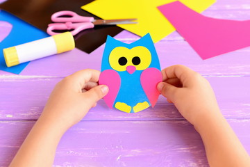 Small child holds paper owl in his hands. Child shows owl crafts. Colored paper sheets, scissors,...