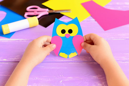 Small child holds paper owl in his hands. Child shows owl crafts. Colored paper sheets, scissors, glue stick on a lilac wooden background. Kids paper crafts. Kids art in kindergarten and at home