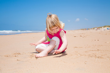 two years old blonde girl with pink dress sitting and writing with finger on golden sand beach with sea behind
