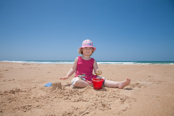 two years old child with pink dress and hat sitting on beach earth ground playing with sand with red bucket and shovels making a castle with ocean behind
