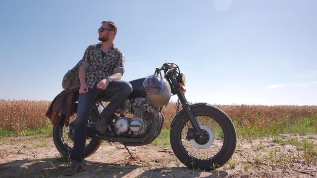 Stylish man smoking on the vintage custom cafe racer in a field.
