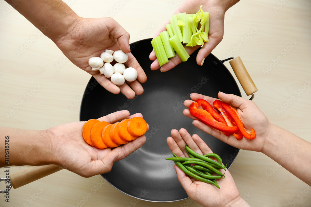 Poster male and female hands holding vegetables above pan on wooden table, top view