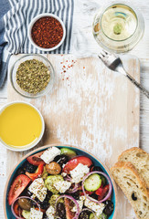Greek salad with olive oil, bread, oregano, pepper and glass of white wine over old white painted wooden board, top view, copy space, verticall composition
