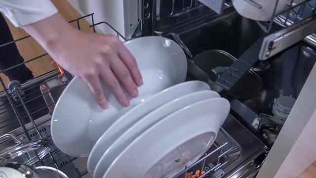 A woman is trying to fill up the dishwasher and she is putting white plates in the lower rank. Close-up shot.
