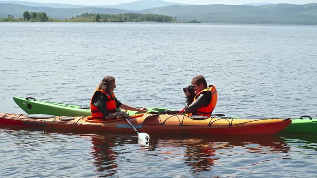 Woman sitting in kayak and holding a paddle while man taking picture of her with professional camera with beautiful lake in the background