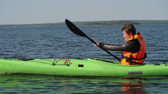 Man in lifejacket and swimsuit sitting in kayak and rowing on the lake with blue water surface 