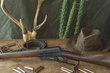 Badezimmer Foto Rückwand Still life. Hunting rifle, antlers, some bullets, vintage trap,belt and cowboy hat on a wooden background in front of hunter clothes © stsvirkun