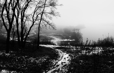Winter landscape with frozen river. Snowy path and trees on top .of hill. Lake is covered with ice in heavy fog. Snowy weather. Black and white photo