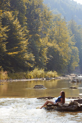 Girl on rocks in river. Beautiful brunette relaxes on shore of mountain river under tall trees. Landscape with mountain river in mist.
