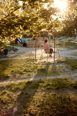 Fototapeta premium Girl on swing. Lonely girl on playground casts long shadow on grass in backlight. Foliage, illuminated by evening sun. Sad mood. loneliness